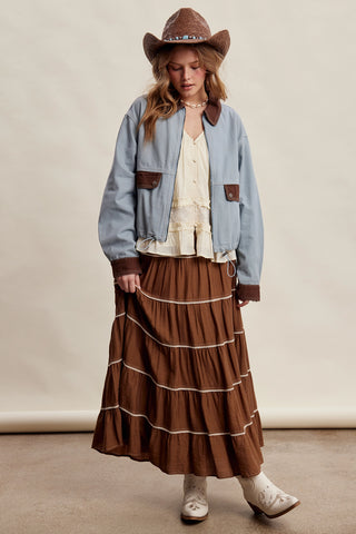 A model wearing a light blue cropped denim jacket with brown pockets, a cream blouse, a brown tiered skirt, a brown cowboy hat, and white cowboy boots, standing against a beige studio backdrop.