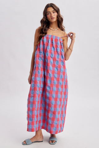 A woman modeling a strapless maxi dress with a pink and blue checkered pattern, paired with light blue slide sandals and a gold necklace, against a plain light gray background.