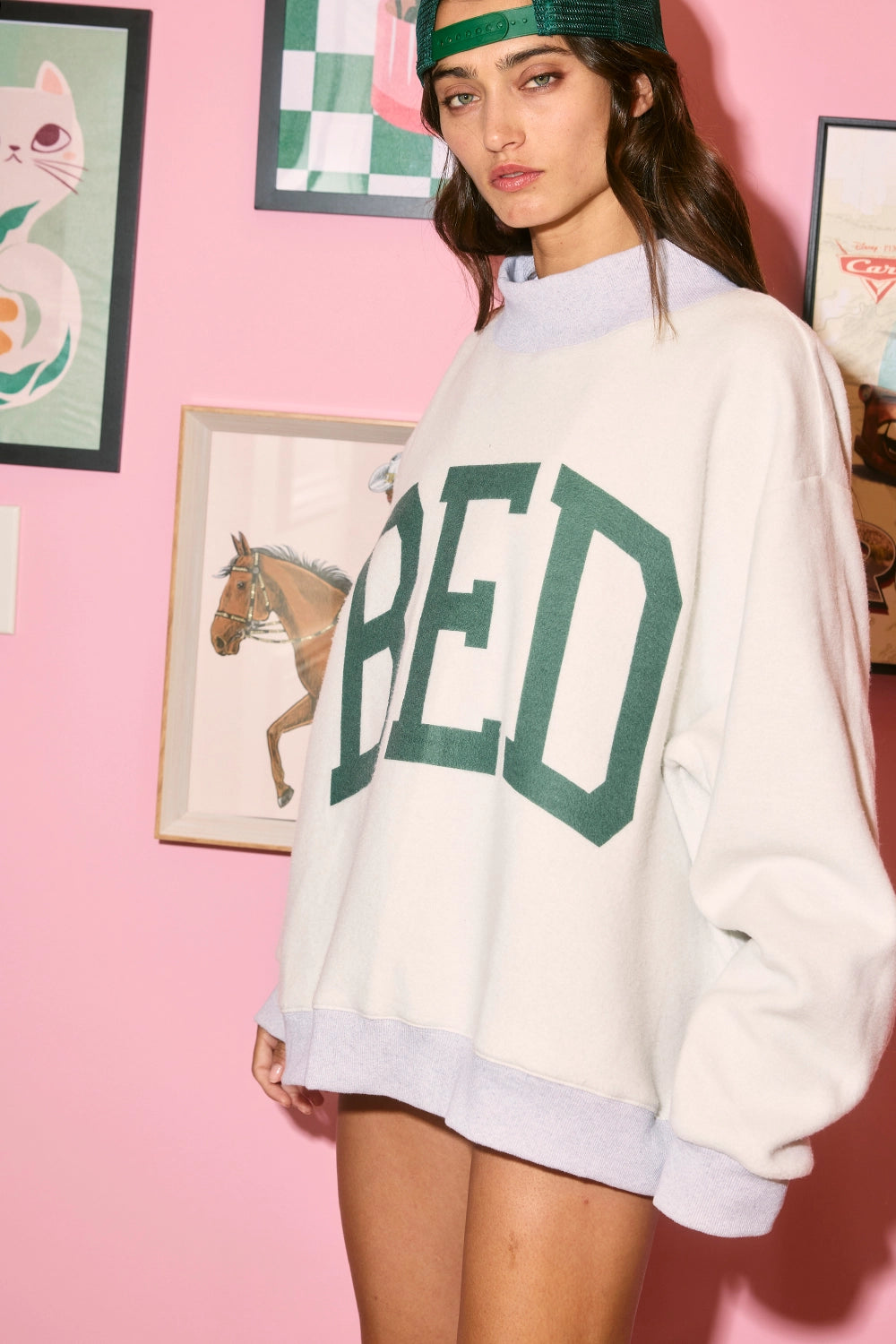 Person wearing a white sweatshirt with green text against a pink wall with framed pictures.