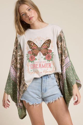 Woman wearing a white t-shirt with a butterfly graphic and text, paired with a green patterned kimono and denim shorts on a beige background.
