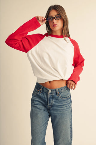 A woman modeling a cropped long-sleeve raglan shirt with a white body and red sleeves, paired with blue jeans, against a plain beige studio background.