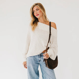 A woman wearing a chunky knit off-the-shoulder cream sweater, light-wash blue jeans, and carrying a dark brown crossbody bag, set against a plain white studio background.