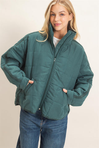 A woman wearing a dark teal-green quilted puffer jacket with a high collar and front zipper, paired with blue jeans, against a plain light beige background.
