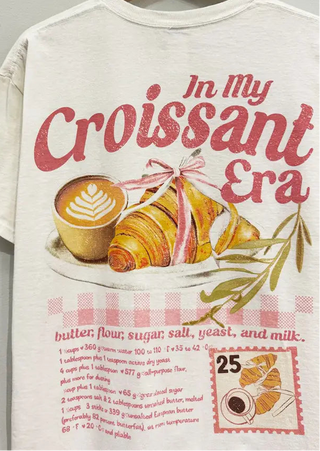 A cream-colored short-sleeve t-shirt hanging on a wooden hanger, featuring a retro-style graphic print with the text 'In My Croissant Era' in red script, an illustration of a croissant tied with a pink ribbon, a cup of latte, and a recipe list below.