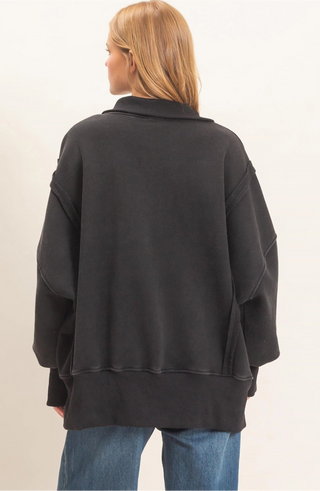 A woman viewed from the back wearing an oversized black long-sleeve top with a wide collar and relaxed fit, paired with blue jeans, against a plain light beige studio background.