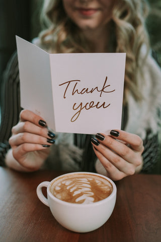 Person holding a 'Thank you' card with a cup of coffee on a wooden table