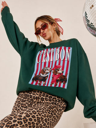 A woman wearing a dark green crewneck sweatshirt with a colorful graphic print of two dachshunds on red and white stripes and the text 'CEST BON'. She is also wearing leopard print pants and sunglasses, posed against a light background with a disco ball partially visible.