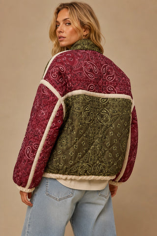 A woman wearing a quilted patchwork jacket with a burgundy paisley pattern, an olive green quilted back panel, and cream shearling trim, shown from the back against a neutral beige background.