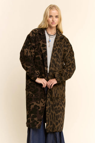 A blonde woman modeling a long brown and black leopard print coat over a light gray top and blue pants, standing against a plain beige studio background.