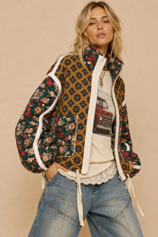 A woman models a patchwork quilted jacket with floral and geometric patterns, featuring cream-colored piping and drawstrings, paired with blue jeans and a graphic t-shirt against a solid beige background.