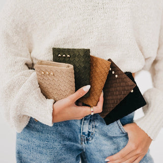 A person in a cream knit sweater and blue jeans holds four small woven zip wallets in beige, olive green, tan, and black, each decorated with small metallic studs.
