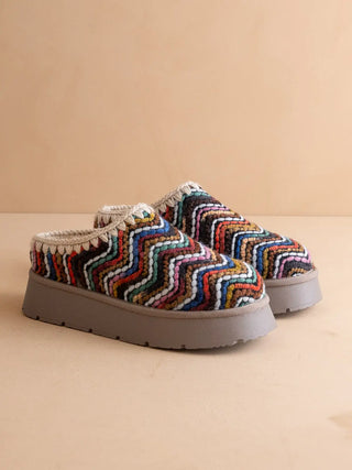A pair of slip-on platform mule shoes featuring a vibrant multicolor chevron knit pattern on the uppers and thick gray platform soles, set against a solid beige background.