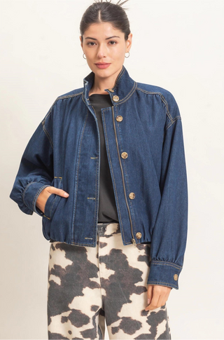 A woman modeling a cropped blue denim jacket with a stand collar and gold-tone buttons, worn over a black top and paired with black-and-white cow-print pants, against a plain light background.