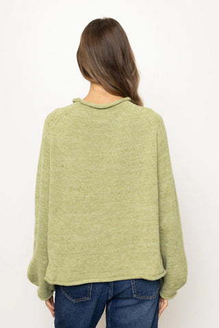 A woman viewed from the back wearing a light olive green knit sweater and blue jeans against a plain white background.