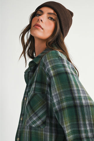 A woman wearing an oversized green and black plaid flannel shirt with a brown knit beanie, photographed against a plain light gray studio background.