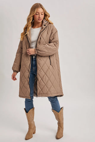 A woman modeling a mid-length quilted puffer coat in taupe brown, styled with a white top, blue jeans, and tan cowboy boots against a light gray studio background.