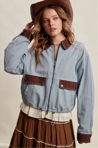 A woman wearing a light blue cropped jacket with brown leather-look trim, collar, cuffs, and chest pockets, paired with a brown hat and a brown pleated skirt, against a plain beige studio background.