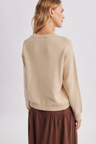 A woman shown from the back wearing a light beige knit sweater with ribbed trim and a brown skirt, photographed against a plain light gray studio background.