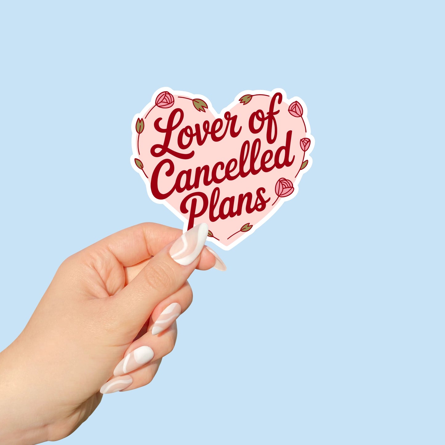Heart-shaped sticker with 'Lover of Cancelled Plans' text held by a hand against a light blue background