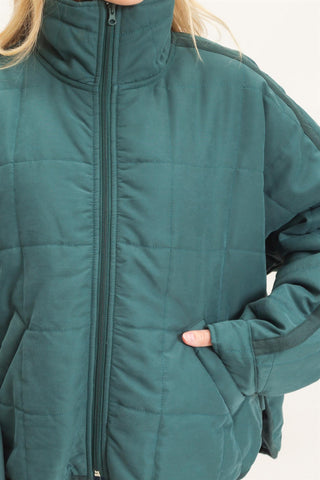 A close-up of a person wearing a dark teal-green quilted puffer jacket with a high collar and front zipper, showing the texture and stitching details.