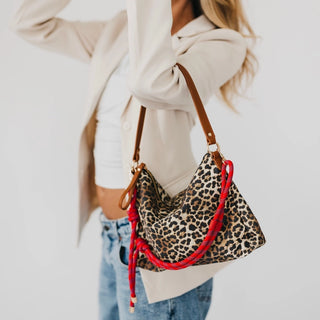 A woman wearing a cream blazer and blue jeans carries a leopard print shoulder bag with a brown leather strap and a bright red braided accent strap against a plain white background.