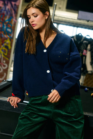 A woman wearing a navy blue knit cardigan with white buttons and green velvet pants, leaning on a pool table in an indoor pool hall setting.