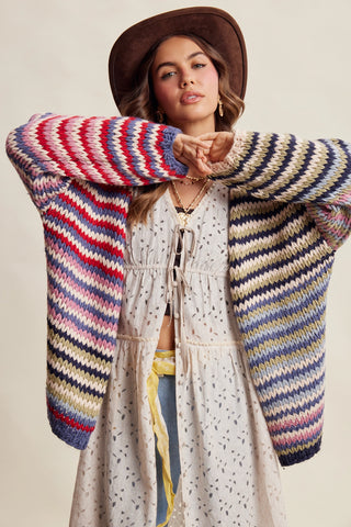 A woman wearing a chunky knit bohemian cardigan with multicolor stripes in red, blue, yellow, green, and cream, styled with a brown wide-brim hat and a cream boho dress.