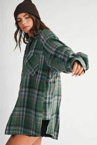 A woman wearing a green plaid flannel shirt dress and a dark brown beanie, posed against a plain light gray studio background.