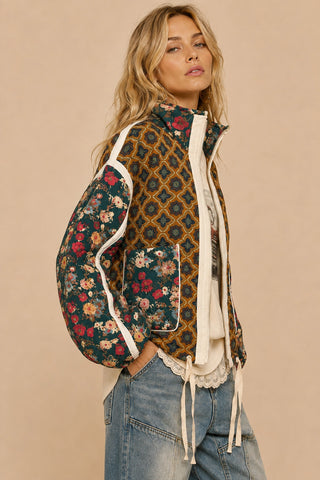 A woman modeling a mixed-media jacket with a dark teal floral pattern on the sleeves and front, a mustard yellow geometric pattern on the back, and white contrast piping, paired with light blue jeans against a solid beige background.