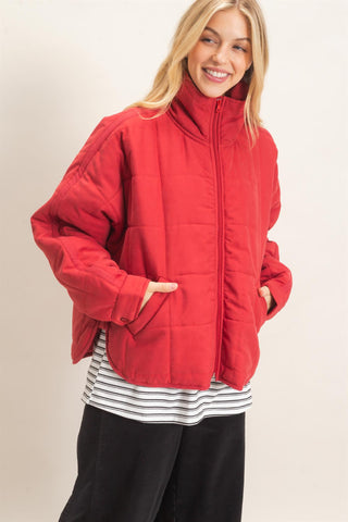 A woman wearing a bright red quilted zip-up jacket over a black and white striped shirt and black pants, standing against a plain light beige studio background.
