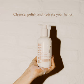 Hand holding a PAUME hand cream bottle with text 'Cleanse, polish and hydrate your hands' on a beige background