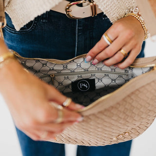 Close-up of a woven beige shoulder bag being opened by a person wearing blue jeans, a cream knit top, and gold jewelry. The bag interior features a black and white monogram pattern and a black brand patch with 'PS' in the center.