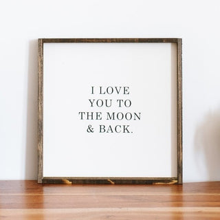 A square wooden framed sign with the text 'I LOVE YOU TO THE MOON & BACK.' centered on it, placed against a white wall on a wooden floor.