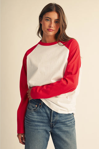 A woman wearing a red and white raglan long-sleeve shirt with blue jeans, standing against a plain beige background.