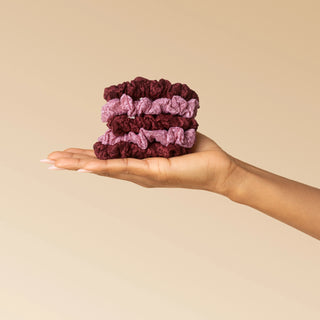A hand holding a small stack of three fabric scrunchies in dark burgundy and light mauve against a plain beige background.