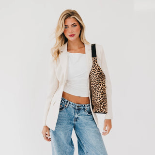 A woman models a leopard print shoulder tote bag with a black strap, styled over a white crop top and blazer with blue jeans against a plain white background.