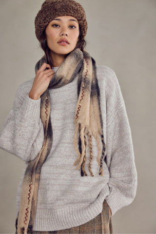 A woman wearing a chunky knit brown beanie, a light gray oversized sweater, and a beige and brown gradient scarf with fringe and zigzag details, posed against a neutral gray studio background.