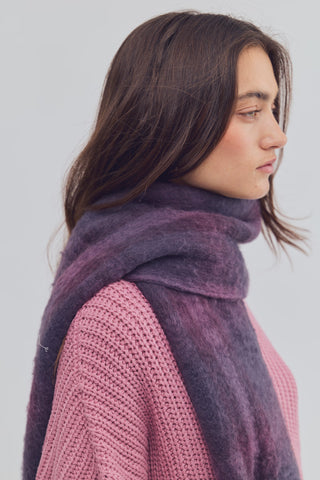 A woman shown in side profile wearing a thick, fuzzy high-neck scarf in a dusty purple shade over a pink knit sweater, against a plain light gray background.