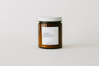 A clear glass jar candle with a white lid, labeled 'ADORE', on a white background.