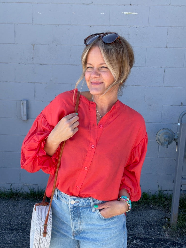 Woman wearing a red shirt and blue jeans standing against a light-colored wall.