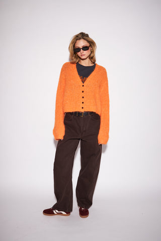 A model wearing a fuzzy orange cardigan over a black top, paired with dark brown wide-leg trousers and burgundy loafers, standing against a plain light gray studio background.