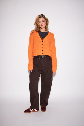A model stands against a light gray studio background wearing a fuzzy orange cropped cardigan over a dark gray top, dark brown corduroy pants, and burgundy sneakers.