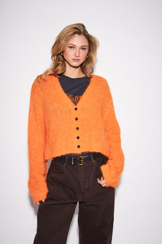 A woman modeling a fuzzy orange cropped cardigan over a black graphic t-shirt and dark brown pants, photographed against a plain light gray studio background.