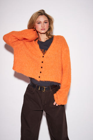 A model wearing a fuzzy, cropped orange cardigan over a dark gray t-shirt and dark brown corduroy pants, posing against a plain white studio background.