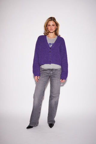 A woman modeling a purple fuzzy button-up cardigan over a gray t-shirt and gray jeans, standing against a light gray studio background.