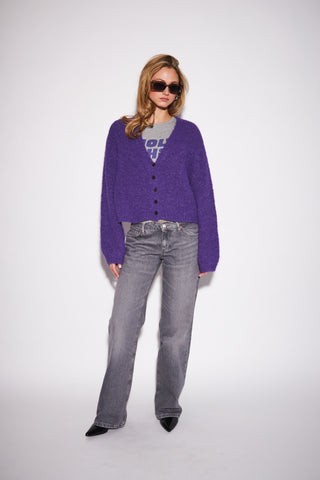 A woman modeling a purple fuzzy button-up cardigan over a gray graphic t-shirt, paired with gray jeans and black pointed shoes, against a plain white studio background.