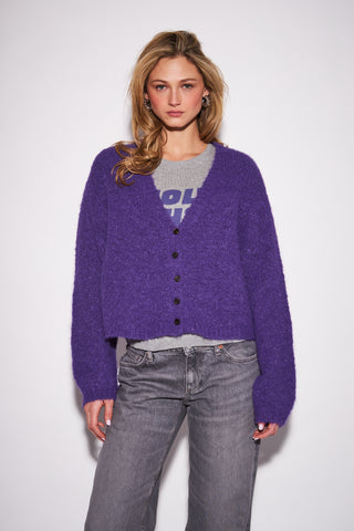 A woman modeling a fuzzy purple button-up cardigan over a gray graphic t-shirt and gray jeans, photographed against a plain light gray studio background.