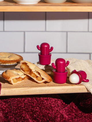 Bright pink cactus-shaped silicone food storage containers in small pots, displayed on a wooden board with apple pie pieces and a white tiled kitchen background.