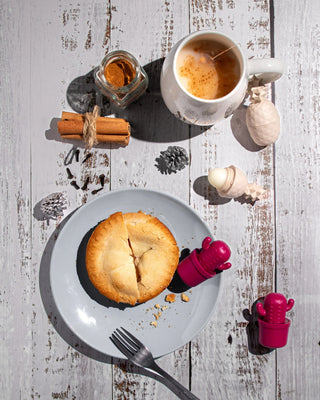 A styled flat-lay of a baked apple pie on a light gray plate, accompanied by a white mug of coffee with milk, cinnamon sticks, a small jar, and decorative objects on a distressed white wooden table.