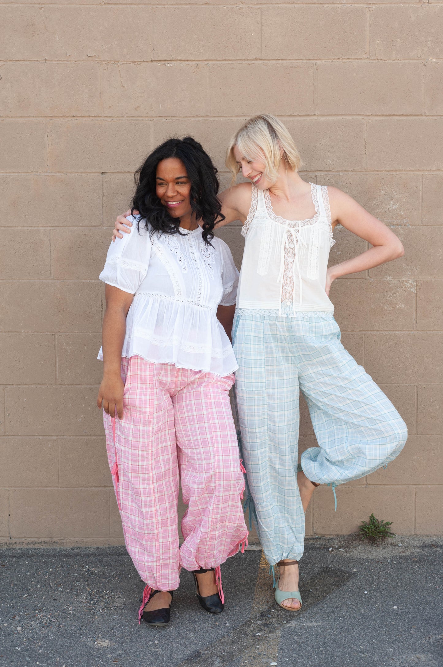 Two women standing against a beige wall, wearing light-colored outfits.